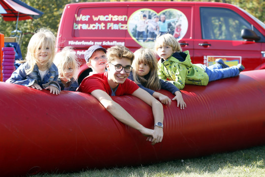 Fünf junge Kinder spielen mit einem Spielbus-Betreuer auf der großen aufblasbaren Luftwurst, hinter ihnen der Spielbus der Wiener Kinderfreunde