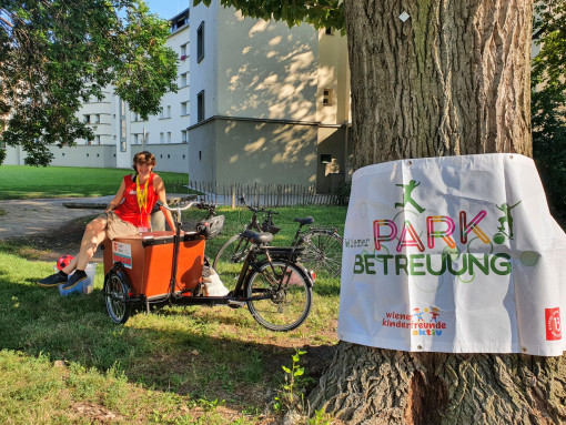 Im Vordergrund ein Baum mit dem Parkbetreuungsplakat, dahinter eine lachende Frau auf einem Lastenfahrrad